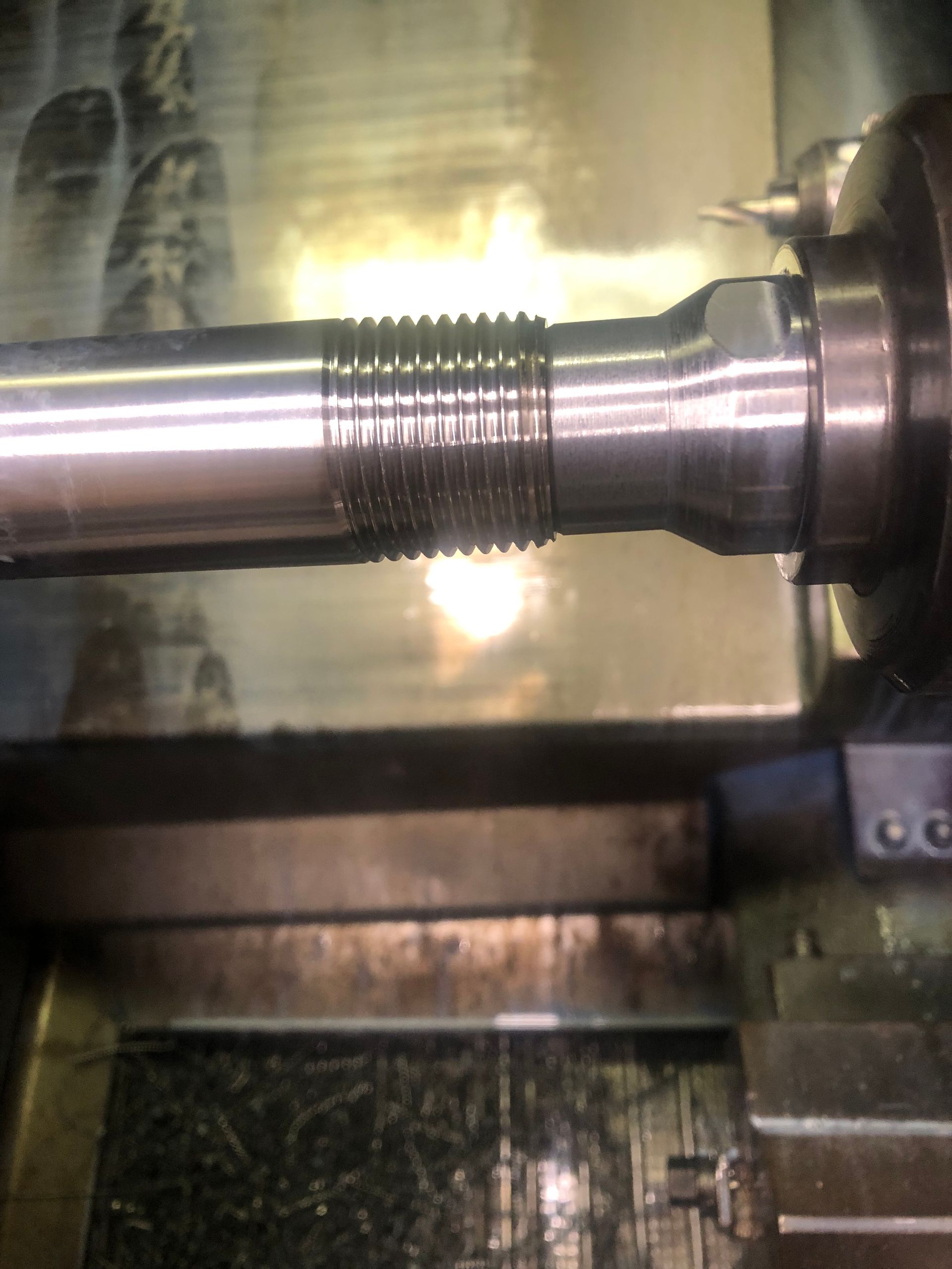 Close-up of a metal shaft in a lathe, with threaded section being machined under bright light