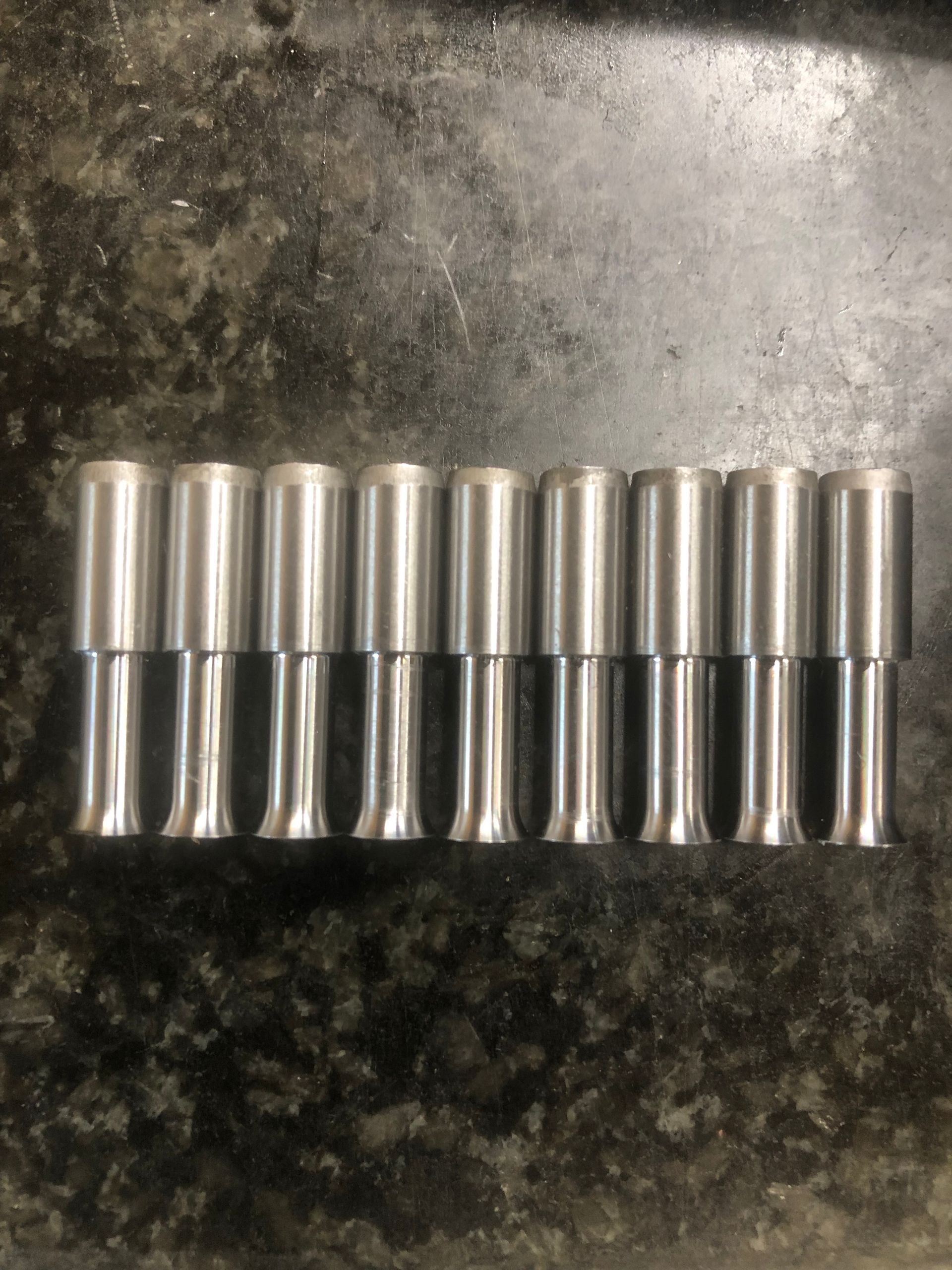 Ten silver cylindrical metal parts lined up on a dark speckled surface