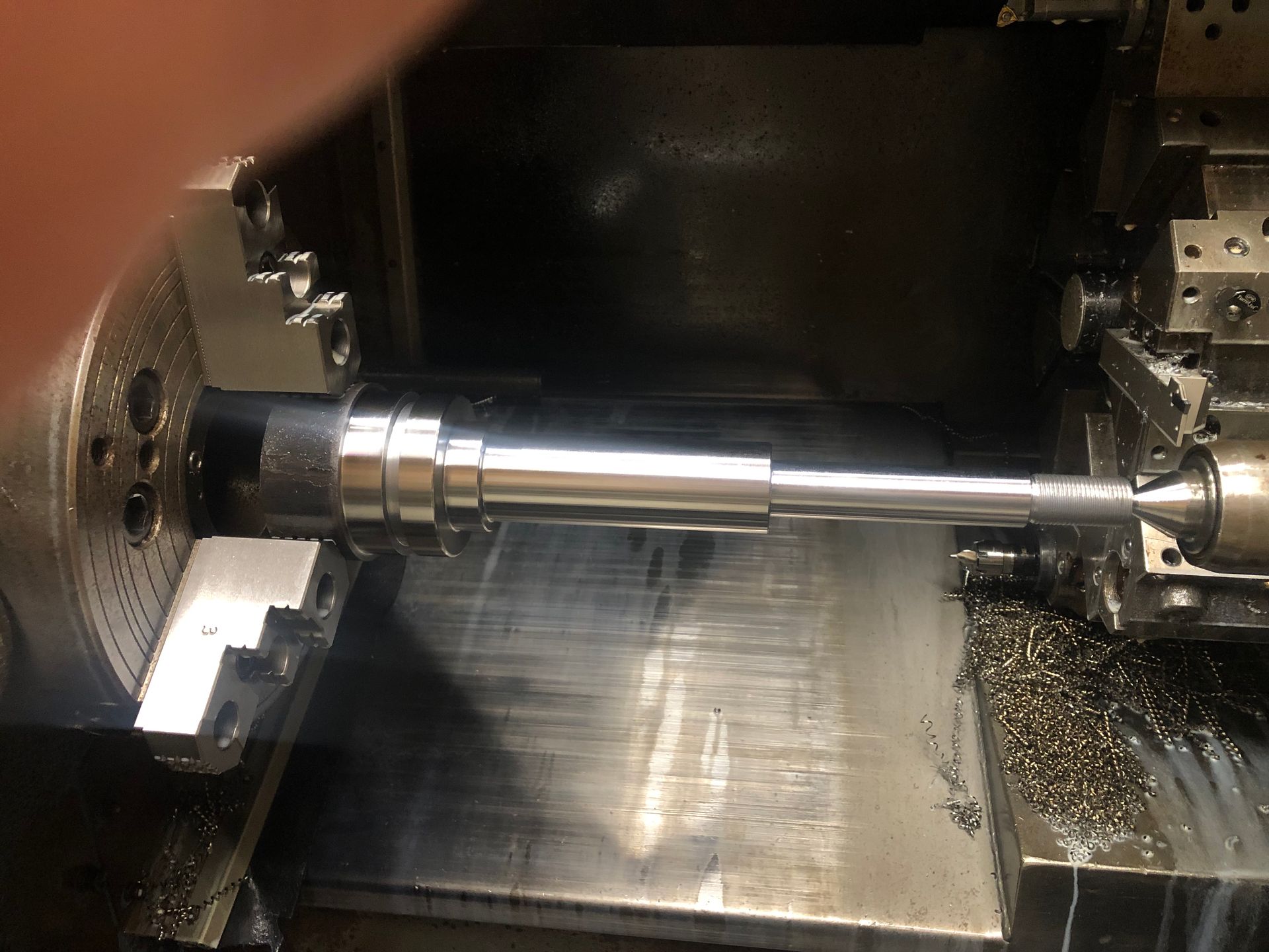 Metal shaft being machined in a lathe, with cutting tools and chips inside the machine.